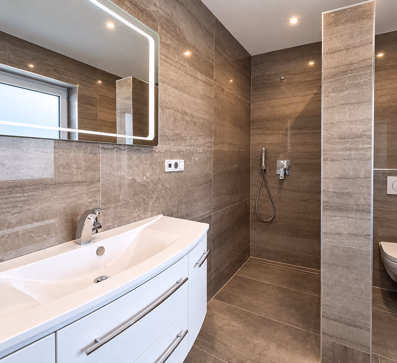 Badezimmer mit grossformatigen Wand- und Bodenfliesen in Marmoroptik, bodengleicher Dusche, Waschtisch und beleuchtetem Spiegel.
