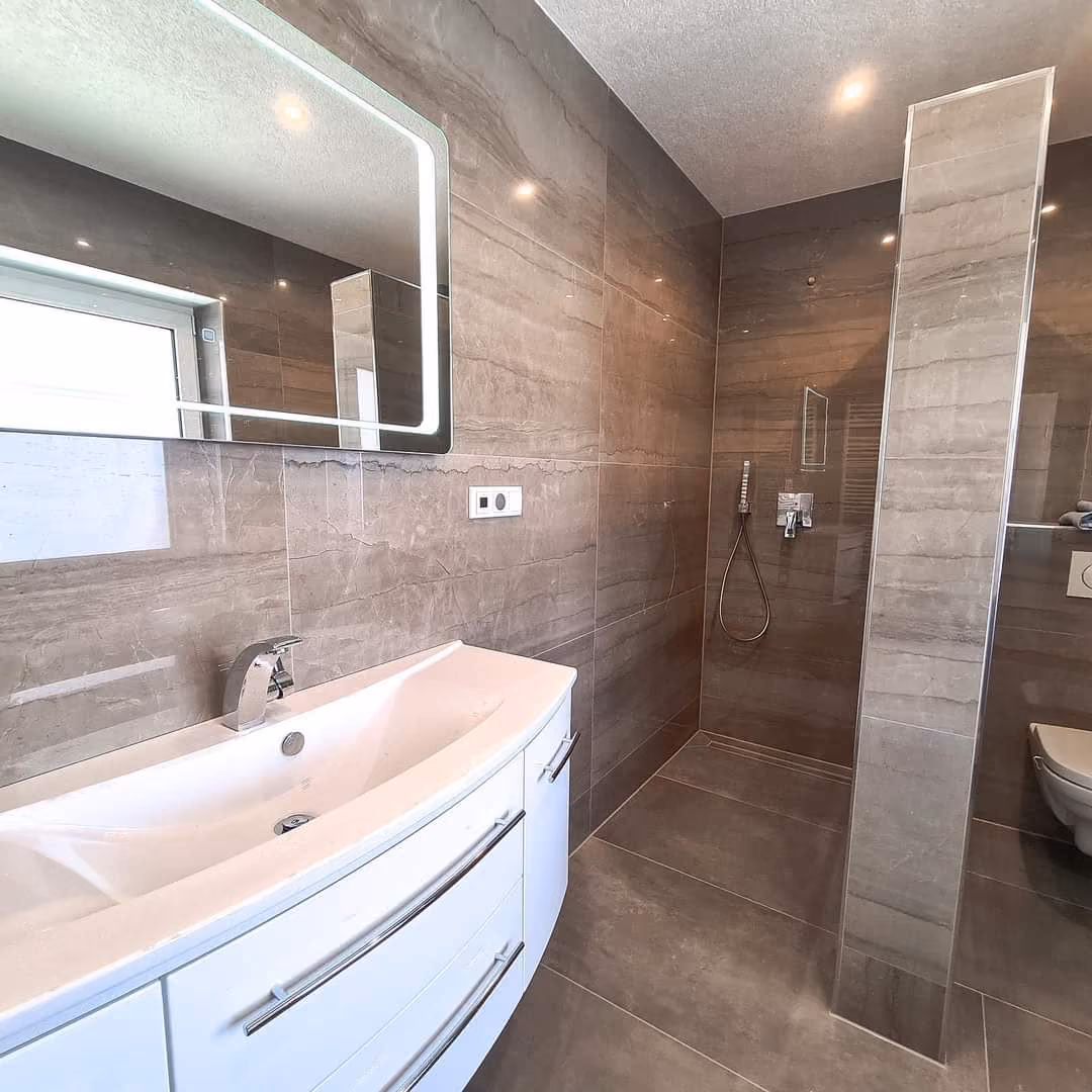 Badezimmer mit braungrauen Wand- und Bodenfliesen, bodengleicher Dusche und beleuchtetem Spiegel ueber dem Waschtisch.